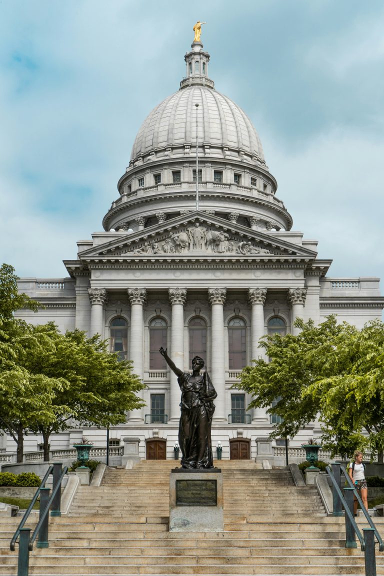 The Wisconsin State Capitol in Madison, viewed head-on up the granite steps. The bronze statue Forward stands at the foot of the columns, her arm raised, beneath the dome and its gilded Wisconsin figure.