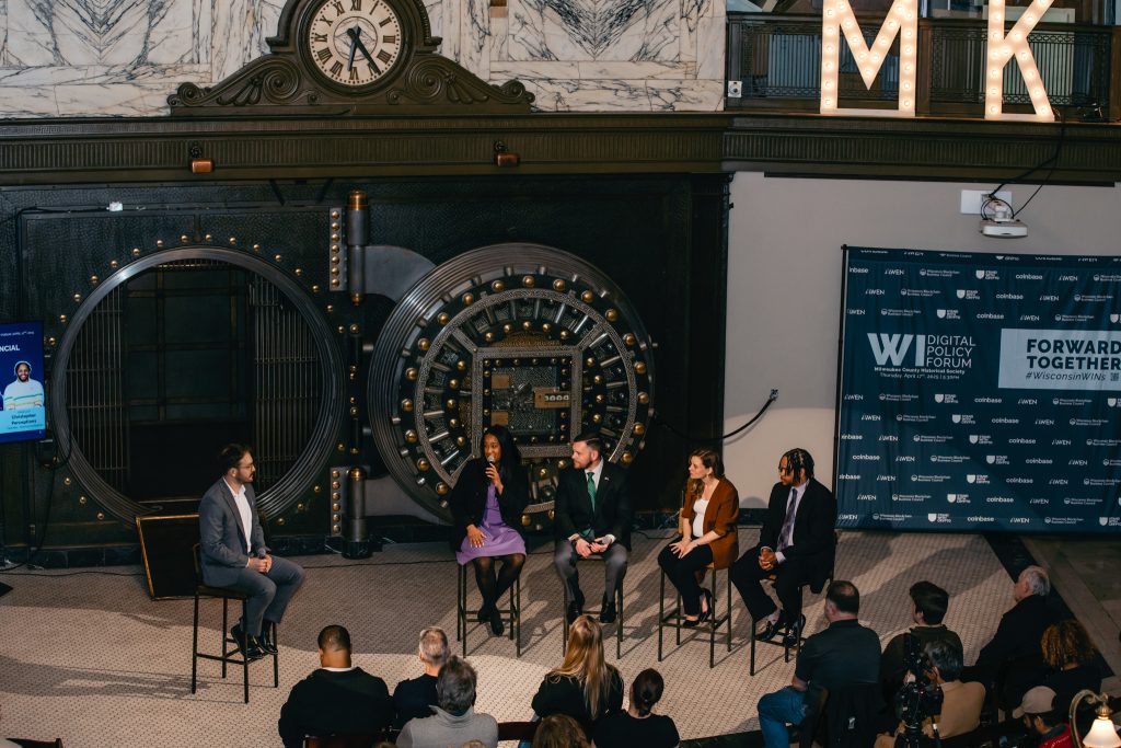 A panel of five seated in front of the building's historic bank vault door, addressing a seated audience.