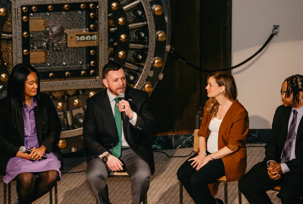 Four panelists seated in conversation beside the open vault door, one gesturing mid-answer.