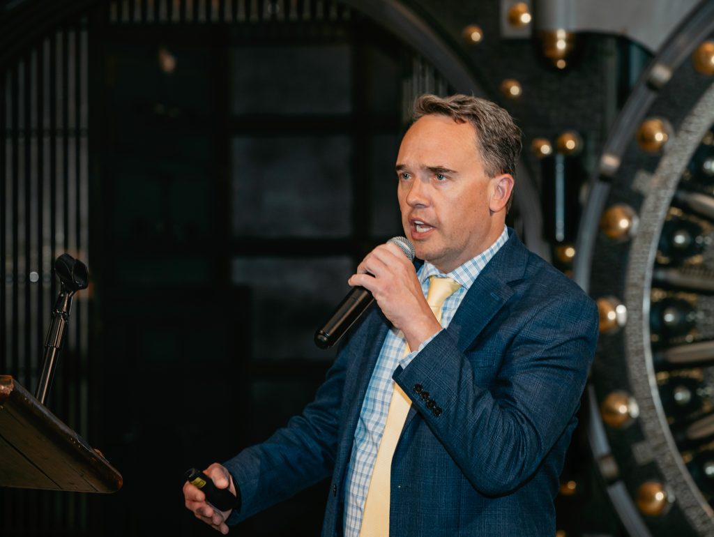 A speaker addressing the room with a handheld microphone against the dark hall.