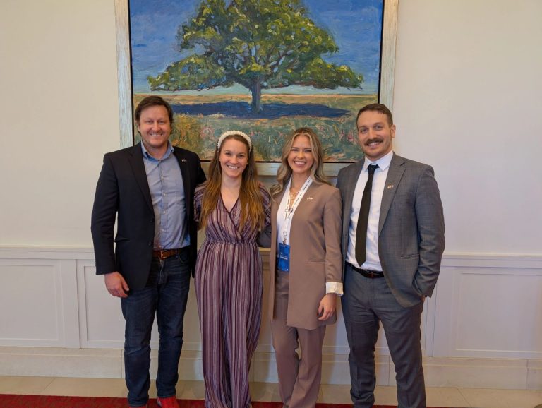 Four members of the WBBC delegation standing together at the North American Blockchain Summit, in front of a large oil painting of a spreading oak tree. One wears a conference lanyard and badge.