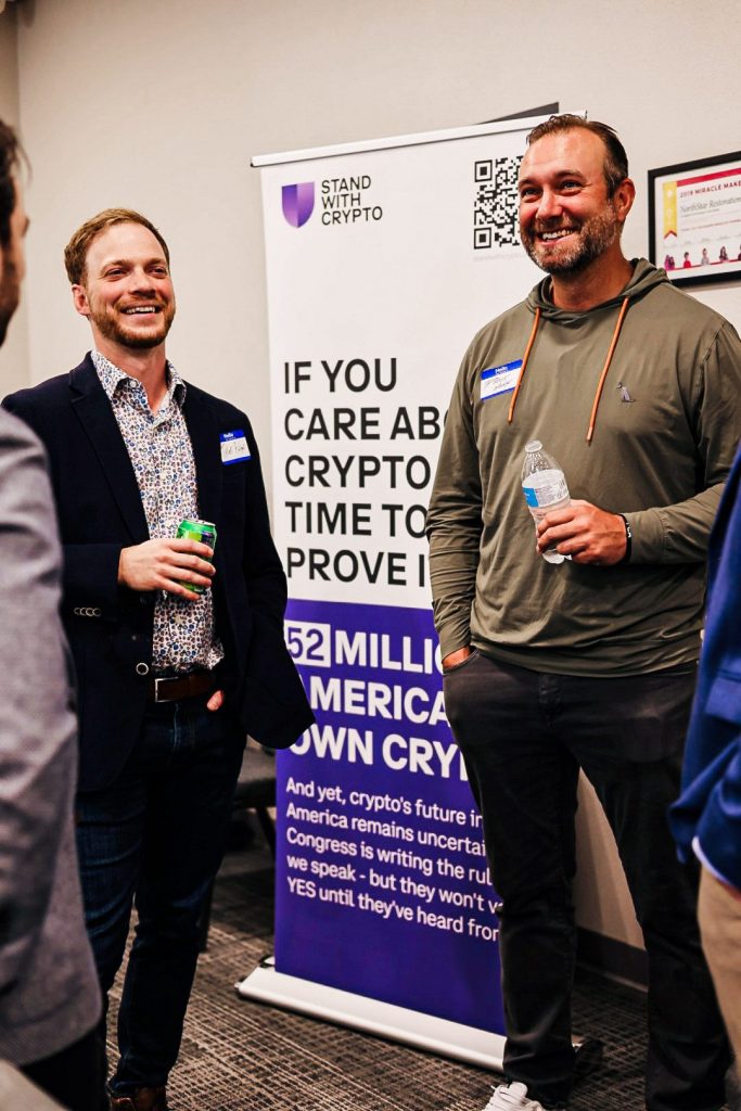 Two attendees talking beside a Stand With Crypto banner at the venue entrance.