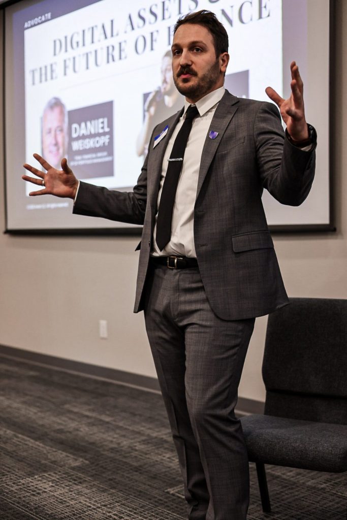 A speaker addressing the room in front of a slide introducing the featured talk on digital assets.