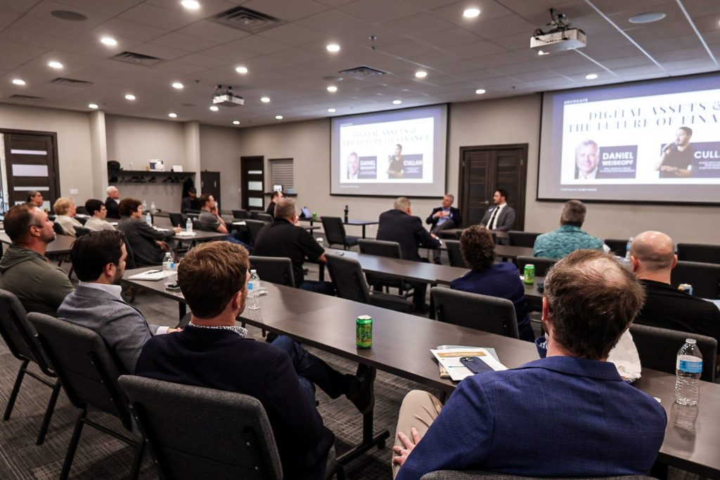 The audience seated in rows watching a presentation, the screen lit at the front of the room.